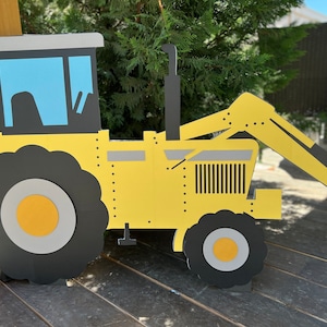 May include: A yellow toy tractor with a black front tire, a black back tire, and a black scoop. The tractor has a gray cab with a blue window.