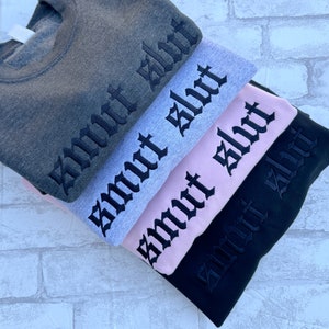 May include: Four sweatshirts stacked on top of each other. The sweatshirts are in different colors: gray, light gray, pink, and black. Each sweatshirt has the word "smut" embroidered on the front in black gothic lettering.