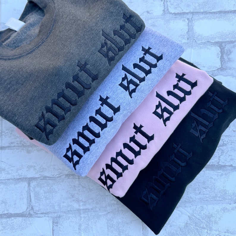May include: Four sweatshirts stacked on top of each other. The sweatshirts are in different colors: gray, light gray, pink, and black. Each sweatshirt has the word "smut" embroidered on the front in black gothic lettering.