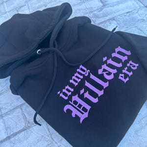 May include: Black hooded sweatshirt with a purple embroidered design that says "in my villain era".