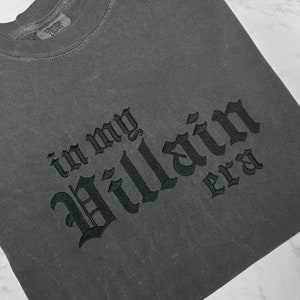 May include: A dark grey t-shirt with the text "in my Villain era" embroidered in black.