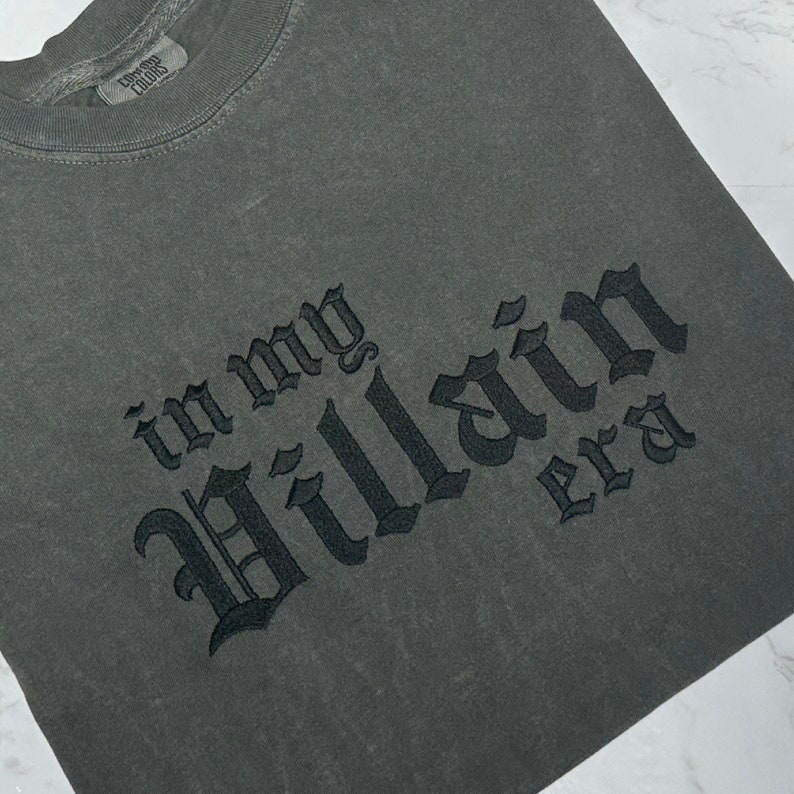 May include: A dark grey t-shirt with the text "in my Villain era" embroidered in black.