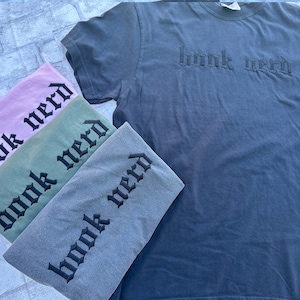 May include: Four t-shirts in different colors, pink, green, gray, and dark blue, with the text "book nerd" in black gothic lettering.