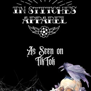May include: A black and white graphic design featuring the text "In Stitches Apparel" and "As Seen on Tik Tok". The design also includes a skull with a crow perched on top, surrounded by flowers and spiderwebs.