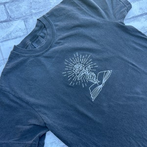 May include: Dark grey t-shirt with a white embroidered design of a rose, hand, and book. The rose is surrounded by radiating lines. The shirt has a crew neck and short sleeves. The brand name "Comfort Colors" is visible on the tag.