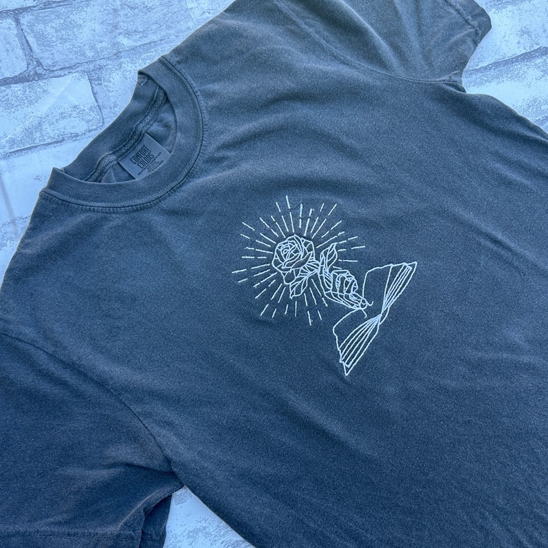 May include: Dark grey t-shirt with a white embroidered design of a rose, hand, and book. The rose is surrounded by radiating lines. The shirt has a crew neck and short sleeves. The brand name "Comfort Colors" is visible on the tag.
