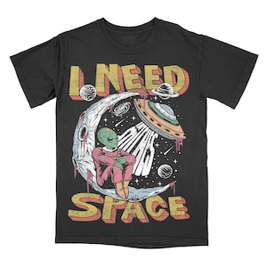 May include: Black t-shirt with a retro graphic featuring an alien seated on a crescent moon, with a UFO and the words "I NEED SPACE". The alien is green and wearing a pink outfit. The design has a vintage, distressed look.