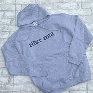 May include: A gray hooded sweatshirt with the text "elder emo" printed in black on the front.