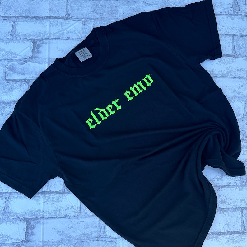 Puede incluir: Camiseta negra con un gr&aacute;fico verde "elder emo" impreso en una fuente g&oacute;tica.