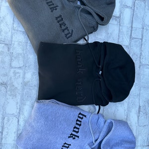 May include: Three hooded sweatshirts in different colors: gray, black, and light gray. Each sweatshirt has the phrase "book nerd" embroidered on the front in black.