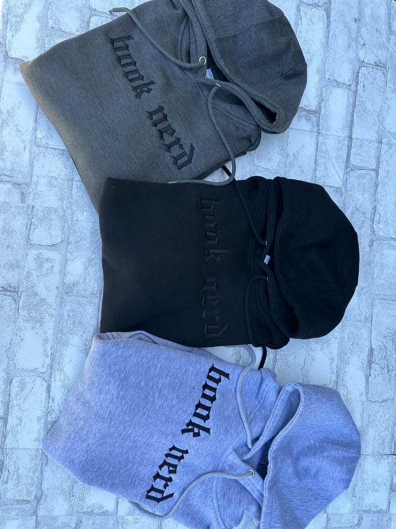 May include: Three hooded sweatshirts in different colors: gray, black, and light gray. Each sweatshirt has the phrase "book nerd" embroidered on the front in black.