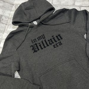 May include: A dark gray hooded sweatshirt with the text "in my Villain era" printed in black on the front.