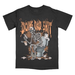 May include: Black t-shirt featuring a skeleton wearing a cap and using a phone while on the toilet. The orange text above reads "DOING DAD SHIT". A humorous design.