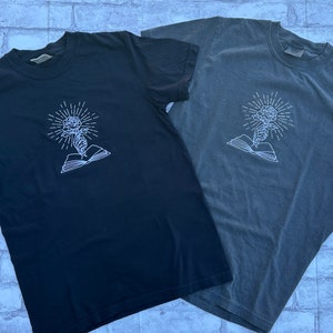 May include: Two short-sleeved t-shirts, one black and one gray, featuring a white embroidered design. The design depicts a rose with radiating lines, emerging from an open book. The shirts are made of a soft material.