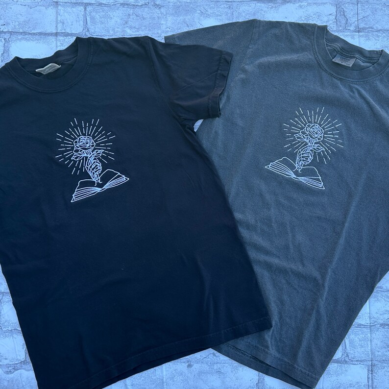 May include: Two short-sleeved t-shirts, one black and one gray, featuring a white embroidered design. The design depicts a rose with radiating lines, emerging from an open book. The shirts are made of a soft material.