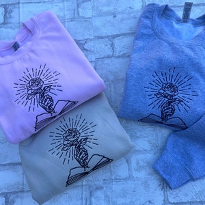 May include: Three sweatshirts in pastel colors: light pink, beige, and gray. Each sweatshirt features a black embroidered design of a rose held by a hand, with radiating lines and an open book.