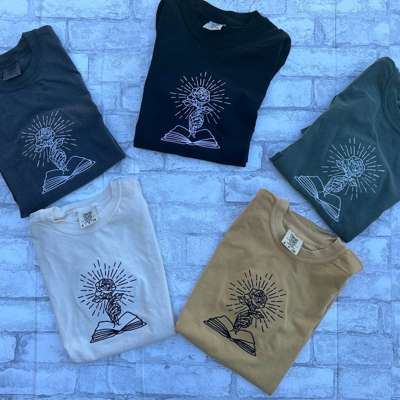 May include: Five t-shirts in various colors: charcoal, black, olive green, cream, and tan. Each shirt features a white graphic of a rose held by a hand emerging from an open book, with radiating lines. The shirts are folded and displayed on a light blue brick surface.