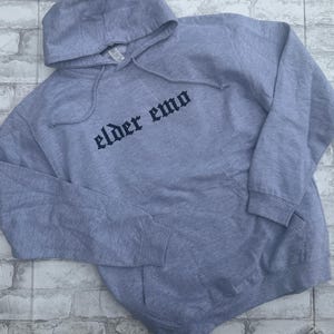 May include: A gray hooded sweatshirt with the text "elder emo" printed in black on the front.
