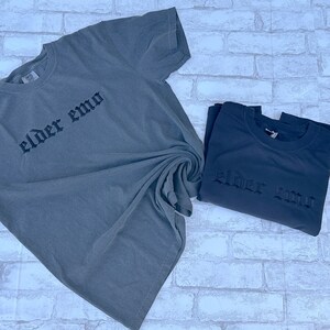 Puede incluir: Dos camisetas con el texto "elder emo" impreso en una fuente g&oacute;tica. Las camisetas son de color gris oscuro y est&aacute;n dobladas.