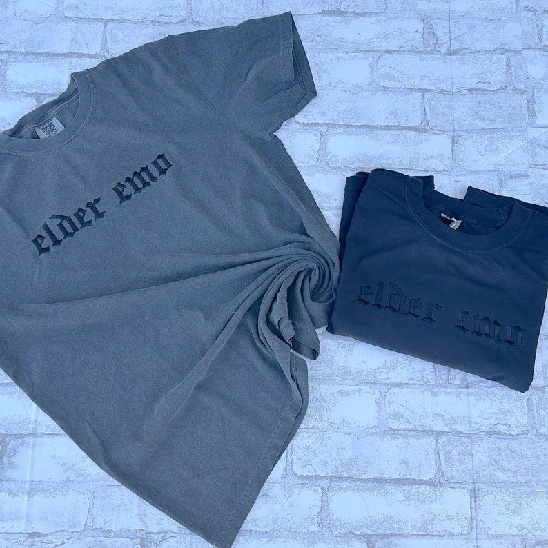 Puede incluir: Dos camisetas con el texto "elder emo" impreso en una fuente g&oacute;tica. Las camisetas son de color gris oscuro y est&aacute;n dobladas.