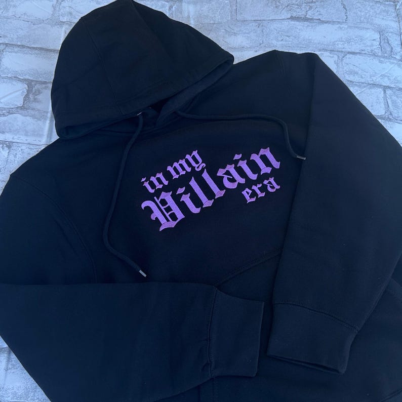 May include: Black hooded sweatshirt with a purple embroidered design that says "in my Villain era".
