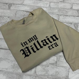 May include: A beige sweatshirt with black embroidered text that reads "in my Villain era".