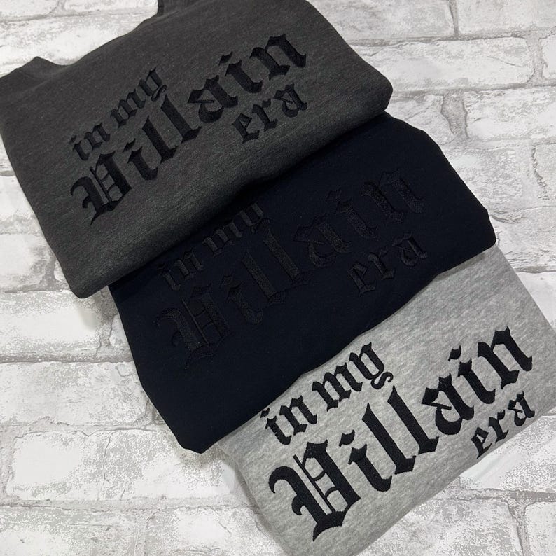 May include: Three sweatshirts stacked on top of each other. The top sweatshirt is dark gray with black embroidered text that reads "in my villain era". The middle sweatshirt is black with black embroidered text that reads "in my villain era". The bottom sweatshirt is light gray with black embroidered text that reads "in my villain era".