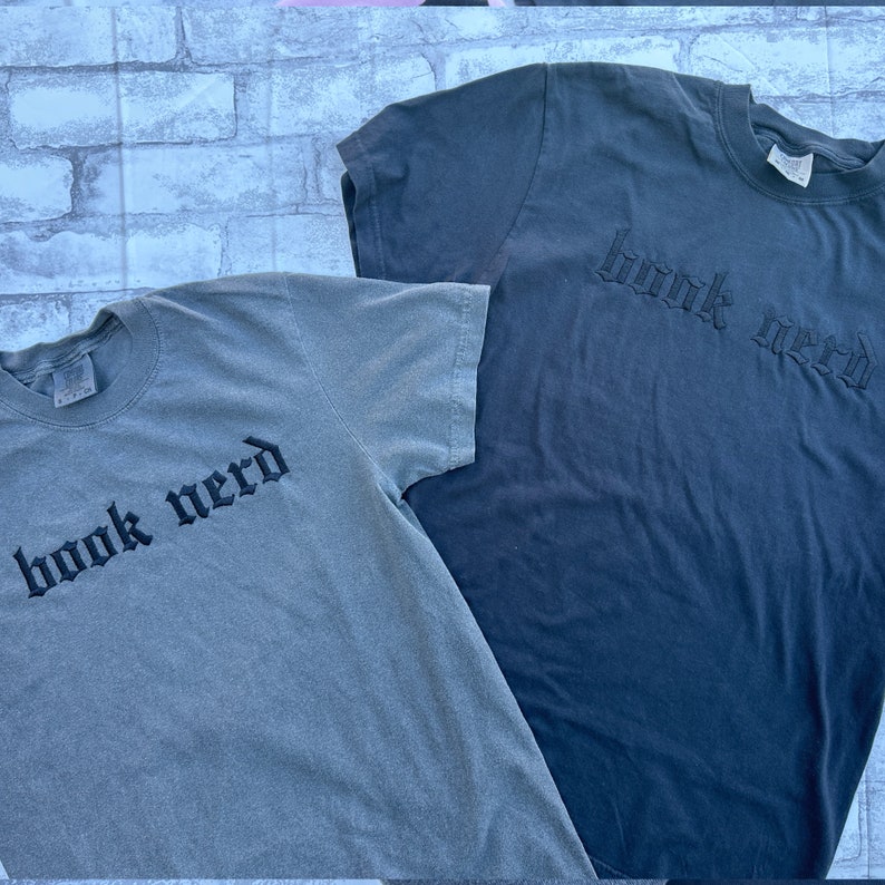 May include: Two t-shirts with the text "book nerd" in a gothic font. One shirt is a dark gray color and the other is a light gray color.