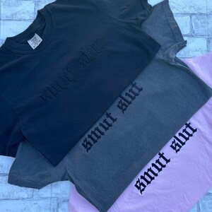 May include: Three t-shirts stacked on top of each other. The top shirt is black with the text "smart shirt" embroidered in black. The middle shirt is gray with the text "smart shirt" embroidered in black. The bottom shirt is pink with the text "smart shirt" embroidered in black.