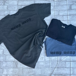 May include: Three t-shirts in different colors, gray, black, and blue, with the same text "WOMP WOMP" in black gothic lettering.