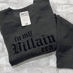 May include: A dark gray sweatshirt with the text "in my Villain era" in black embroidered lettering.