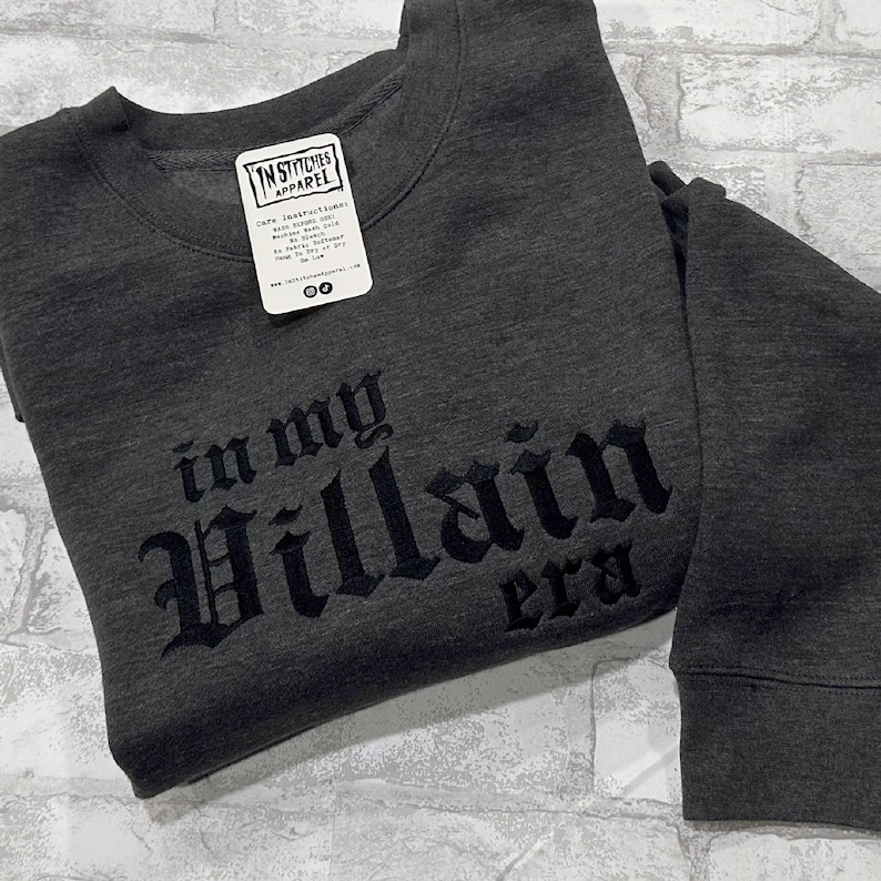 May include: A dark gray sweatshirt with the text "in my Villain era" in black embroidered lettering.