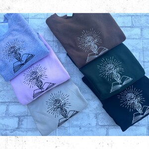 May include: A stack of sweatshirts in various colors, including gray, brown, green, and black. Each sweatshirt features an embroidered design of a rose held by a hand emerging from an open book, with radiating lines.