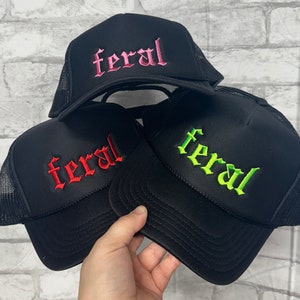 May include: Three black trucker hats with mesh back panels. The hats have the word "feral" embroidered on the front in different colors: pink, red, and green.