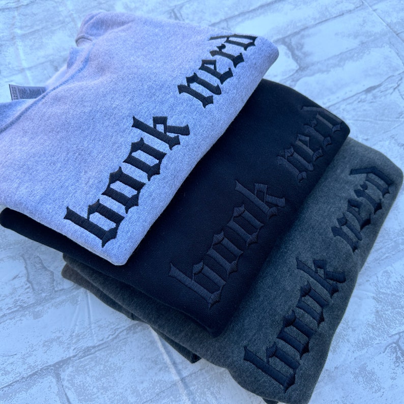 May include: Three sweatshirts stacked on top of each other. The sweatshirts are gray, black, and dark gray. Each sweatshirt has the words "book nerd" embroidered on the sleeve in a gothic font.