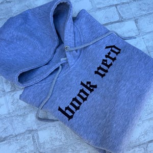 May include: A heather gray hooded sweatshirt featuring a drawstring hood and the text "book nerd" printed in a bold, black, gothic-style font across the front. The garment is made of a soft, textured fabric with visible gray and white fibers.