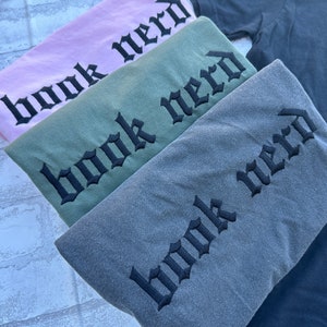 May include: Four t-shirts stacked on top of each other. The shirts are in different colors: pink, green, gray, and black. Each shirt has the words "book nerd" embroidered on the front in black gothic lettering.
