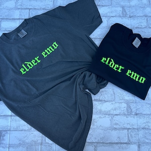 Puede incluir: Dos camisetas con el texto "elder emo" en letras g&oacute;ticas verdes. Las camisetas son una gris oscuro y otra negra.
