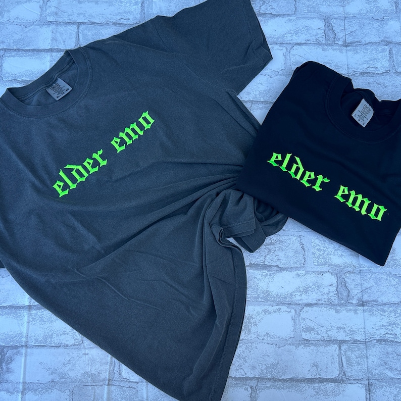 Puede incluir: Dos camisetas con el texto "elder emo" en letras g&oacute;ticas verdes. Las camisetas son una gris oscuro y otra negra.