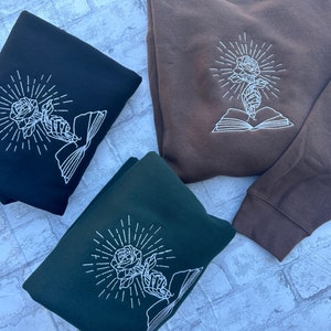 May include: Three sweatshirts in black, brown, and dark green, each featuring a white embroidered design. The design depicts a rose held by a hand, radiating light, and emerging from an open book. The sweatshirts are folded, showcasing the design prominently.