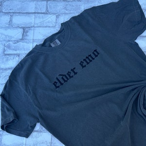 Puede incluir: Una camiseta gris oscuro con las palabras "elder emo" impresas en letras g&oacute;ticas negras.