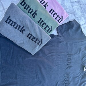 May include: Four t-shirts stacked on top of each other. The shirts are in different colors: gray, green, pink, and dark blue. Each shirt has the words "book nerd" embroidered on the front in a gothic font.