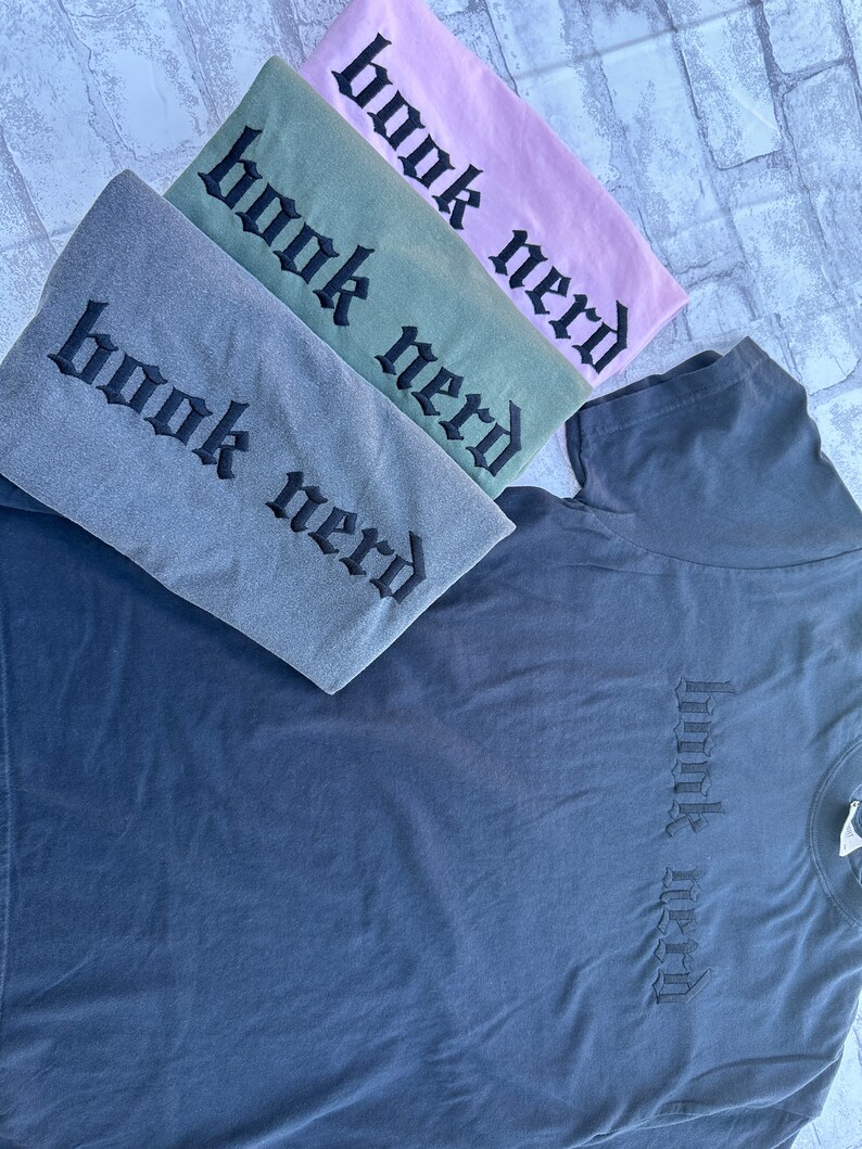May include: Four t-shirts stacked on top of each other. The shirts are in different colors: gray, green, pink, and dark blue. Each shirt has the words "book nerd" embroidered on the front in a gothic font.
