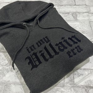 May include: A dark gray hooded sweatshirt with the text "In my Villain era" printed in black.