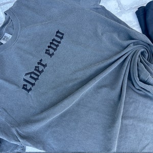 May include: A gray t-shirt with the text "elder emo" embroidered in black on the front.