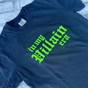 May include: Black t-shirt with green embroidered text that reads "in my villain era".