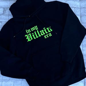 May include: Black hooded sweatshirt with a green embroidered design that says "in my Villain era".