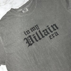 May include: A grey t-shirt with the text "in my Villain era" embroidered in black.