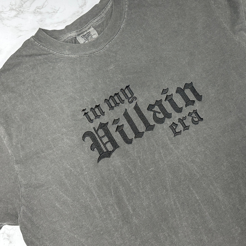 May include: A grey t-shirt with the text "in my Villain era" embroidered in black.