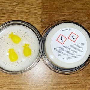 Könnte beinhalten: Ein weißes Kerzengefäß mit einem durchsichtigen Deckel. Das Gefäß enthält eine weiße Kerze mit drei gelben, ananasförmigen Wachsverzierungen. Das Gefäß trägt ein Etikett mit der Aufschrift "Pineapple" und enthält einen Warnhinweis auf mögliche allergische Reaktionen und Toxizität.